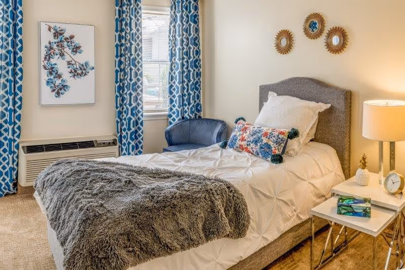 A cozy bedroom with a single bed featuring a gray upholstered headboard, white bedding, a colorful decorative pillow, and a gray furry throw blanket. Next to the bed is a white nightstand with a lamp, a small clock, a pineapple decoration, and a tissue box. A blue armchair is placed near a window with blue and white patterned curtains. The walls are light-colored with three decorative round mirrors and a framed artwork of blue flowers.