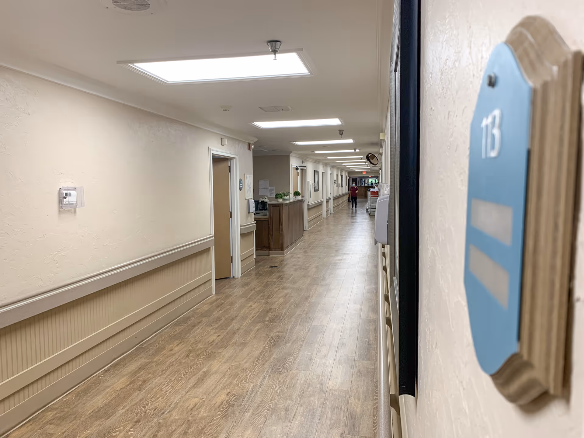 A long, well-lit hallway in a healthcare facility with wood-patterned flooring and beige walls. Room number 113 is visible on a blue plaque on the right side of the image. A person is seen walking in the distance near the end of the hallway, and there is a reception or nurse station along the left side.