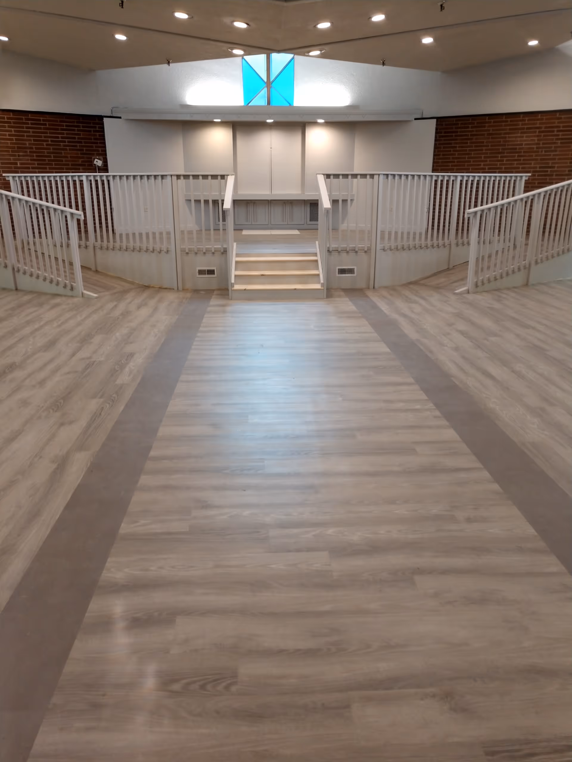 Interior view of a spacious room with light wood flooring and a raised platform area with white railings and steps in the center. The walls are a combination of white and brick, and there is a window with blue and white stained glass near the ceiling.