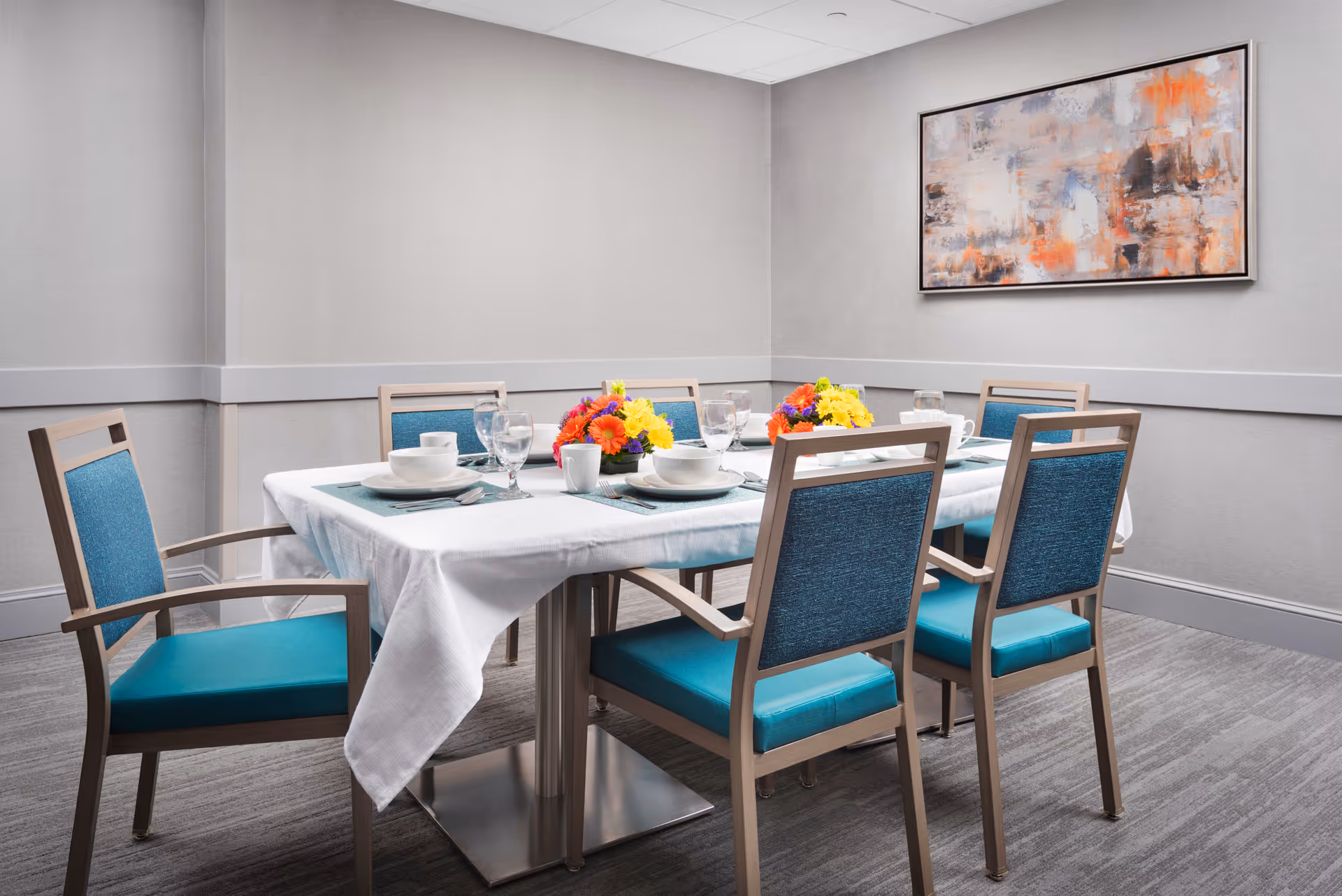 A dining room with a rectangular table covered with a white tablecloth. Six wooden chairs with teal cushions surround the table. The table is set with white plates, bowls, cups, and glassware, along with teal placemats and silverware. Two colorful floral centerpieces are placed on the table. The room has light gray walls, a gray carpet, and a large abstract painting with orange, gray, and white tones hanging on the wall.