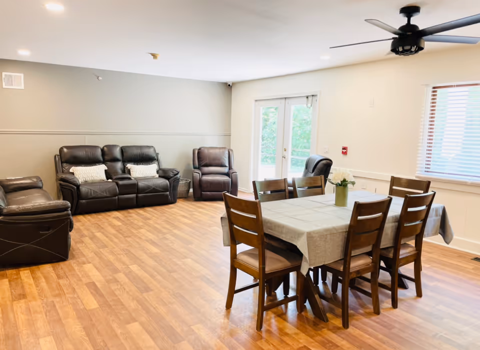 A bright and spacious common area featuring a wooden dining table with six chairs covered by a beige tablecloth and a vase with white flowers. To the left, there are three dark brown leather recliners and a matching loveseat arranged against a light gray wall. The room has wood flooring, a ceiling fan, recessed lighting, a window with blinds, and glass double doors leading outside.