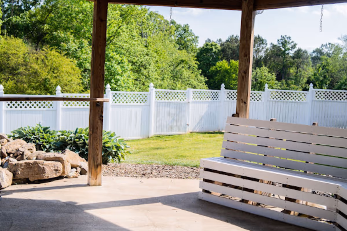 A covered outdoor patio area with a white wooden bench swing, wooden posts supporting the roof, a white fence in the background, green grass, and trees beyond the fence.