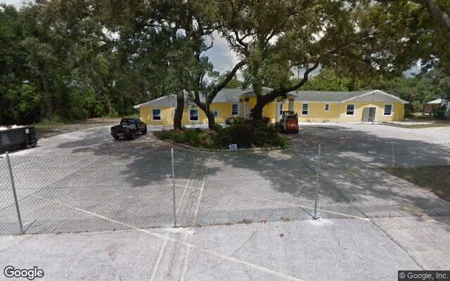 Exterior view of a single-story yellow building with a white roof, surrounded by trees and a chain-link fence in the foreground. There are a few vehicles parked near the building and a small landscaped area with large trees in front.