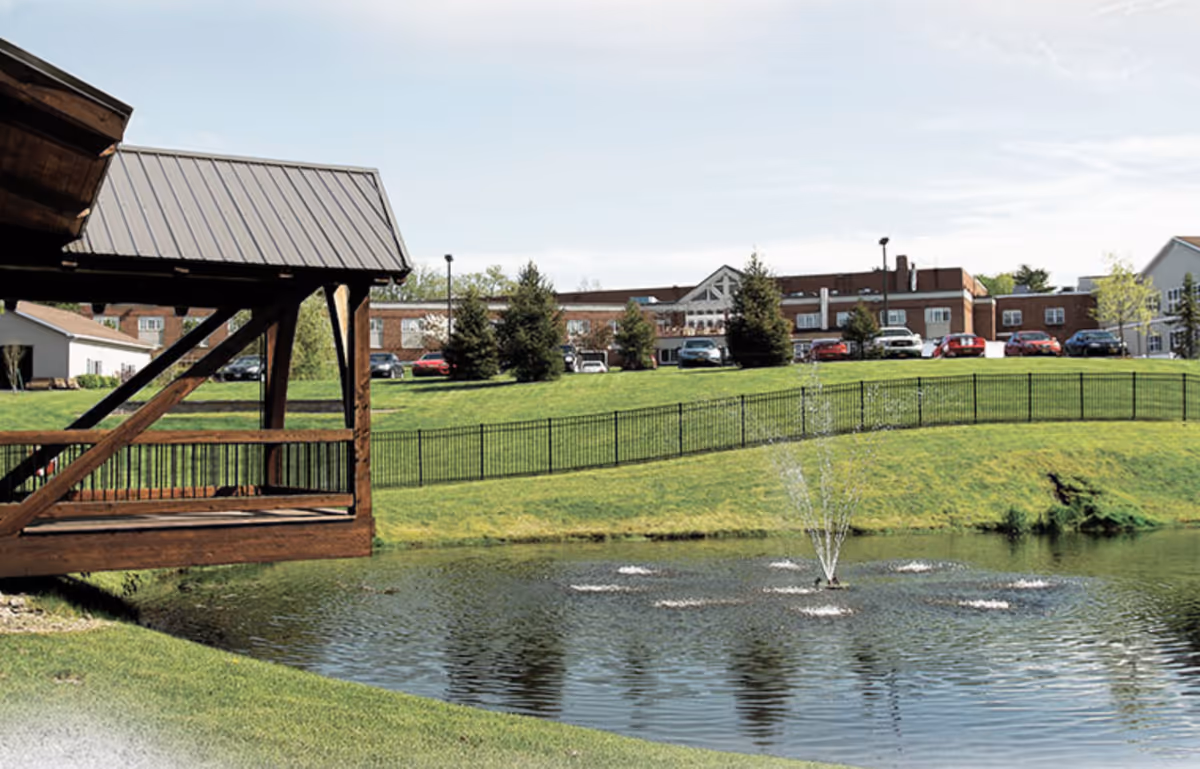 A peaceful outdoor scene at Tanglewood Manor featuring a small pond with a water fountain in the center, a wooden covered bridge on the left, green grassy hills, a black metal fence, several parked cars, and a large brick building in the background under a clear sky.