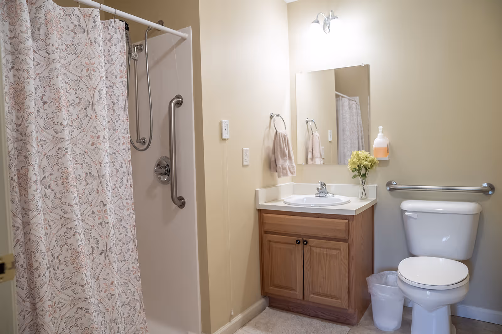 A clean bathroom featuring a shower with a patterned curtain and a grab bar, a wooden vanity with a sink and mirror above it, a towel hanging on a ring, a small vase with flowers on the counter, a wall-mounted soap dispenser, a toilet with a grab bar behind it, and a small trash can on the floor.