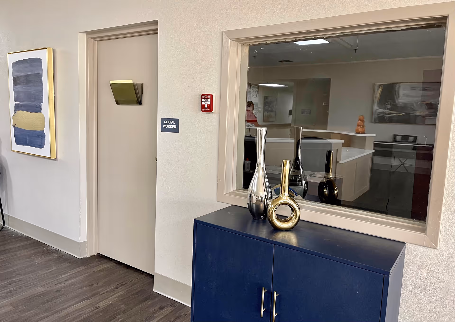 Interior hallway area in a nursing and rehabilitation facility with a closed door labeled 'Social Worker'. Next to the door is a large window looking into an office space. Below the window is a dark blue cabinet with three decorative vases in silver, gold, and black colors. A modern abstract painting hangs on the wall to the left of the door.