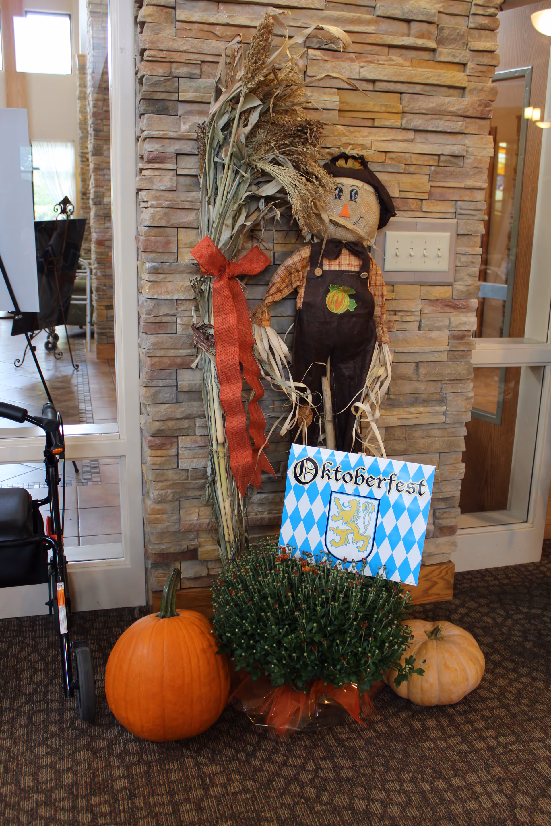 Seasonal autumn display with pumpkins, a potted mum, a scarecrow and an "Oktoberfest" sign against a stone interior wall.