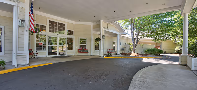 Covered drive-through entrance with automatic glass doors, benches, an American flag, and landscaped grounds at an assisted living facility.