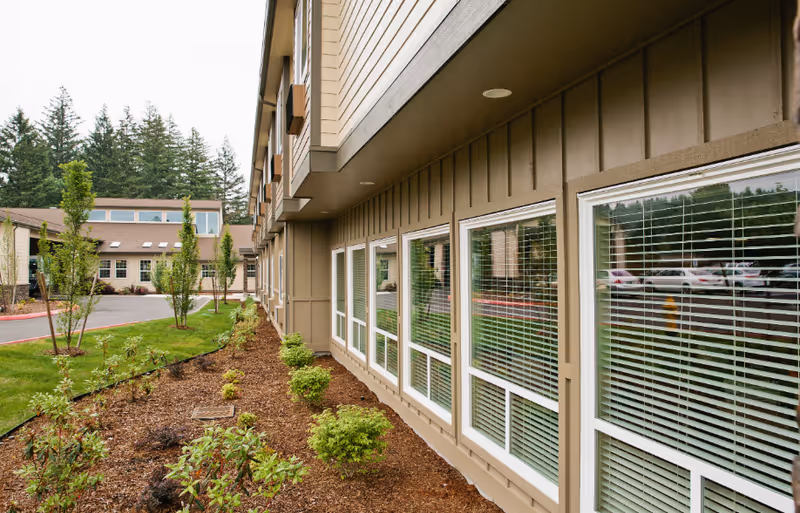 Exterior view of a senior living facility showing a landscaped garden with small bushes and young trees along the side of a beige building with multiple windows featuring white blinds. A parking area with cars and tall evergreen trees are visible in the background.
