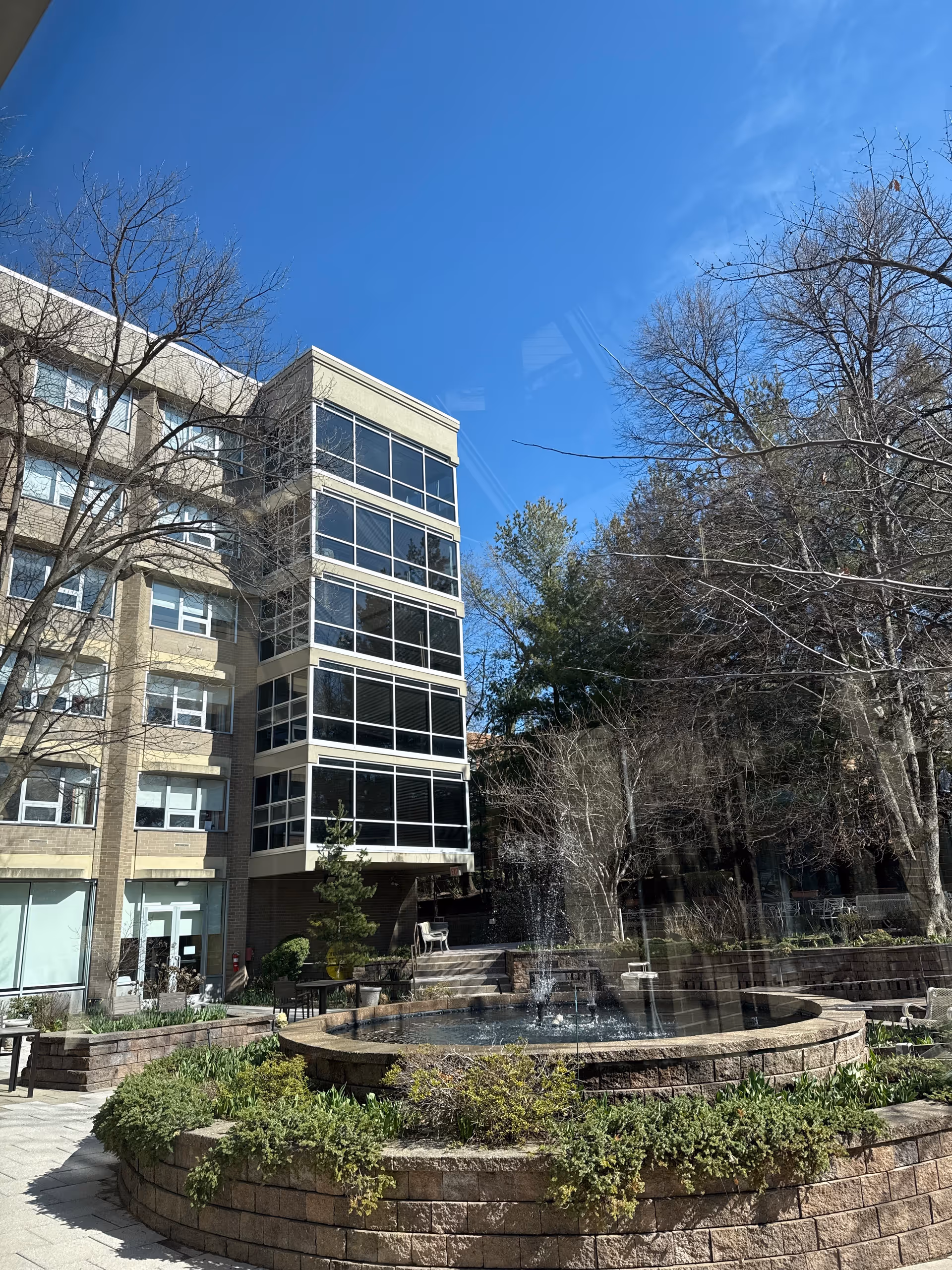 Outdoor courtyard area of Margaret Tietz Nursing and Rehabilitation Center featuring a circular stone fountain with water jets, surrounded by greenery and trees with bare branches. The multi-story building with large windows is visible in the background under a clear blue sky.