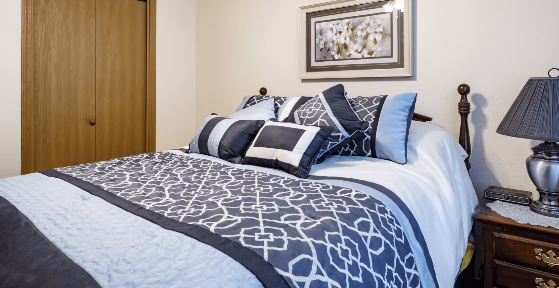 A neatly made bed with a blue and white patterned comforter and multiple matching pillows in a bedroom. There is a wooden nightstand with a lamp and an alarm clock next to the bed, and a framed floral painting hangs on the wall above the bed. A wooden closet door is visible in the background.