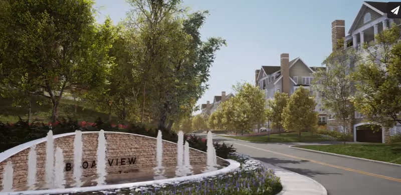 A landscaped entrance area with a curved stone wall featuring water fountains and the partially visible text 'BROADVIEW'. The scene includes a paved road lined with trees and multi-story residential buildings in the background under a clear sky.
