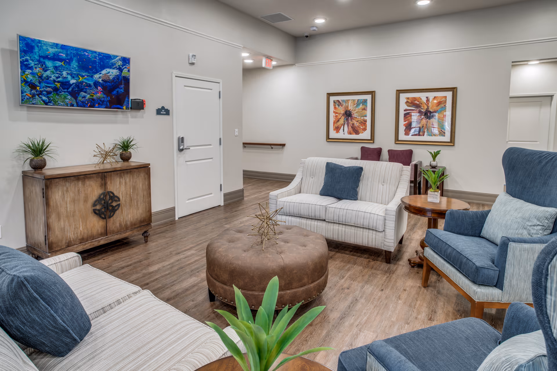A cozy living room area in a senior living facility featuring two striped sofas with blue cushions, two blue armchairs, a round brown ottoman in the center, and a wooden cabinet with decorative plants. The walls are adorned with two colorful abstract paintings and a large aquarium mounted above the cabinet. The room has wooden flooring and soft lighting.