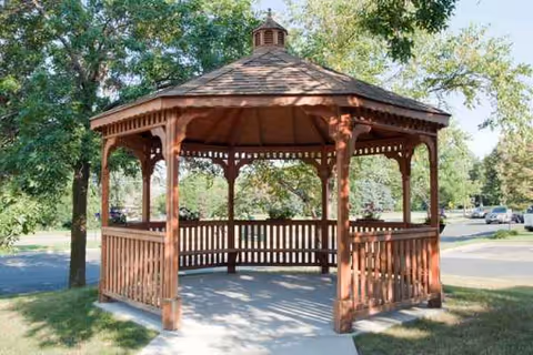 Wooden gazebo in a sunny outdoor park with trees and a sidewalk.