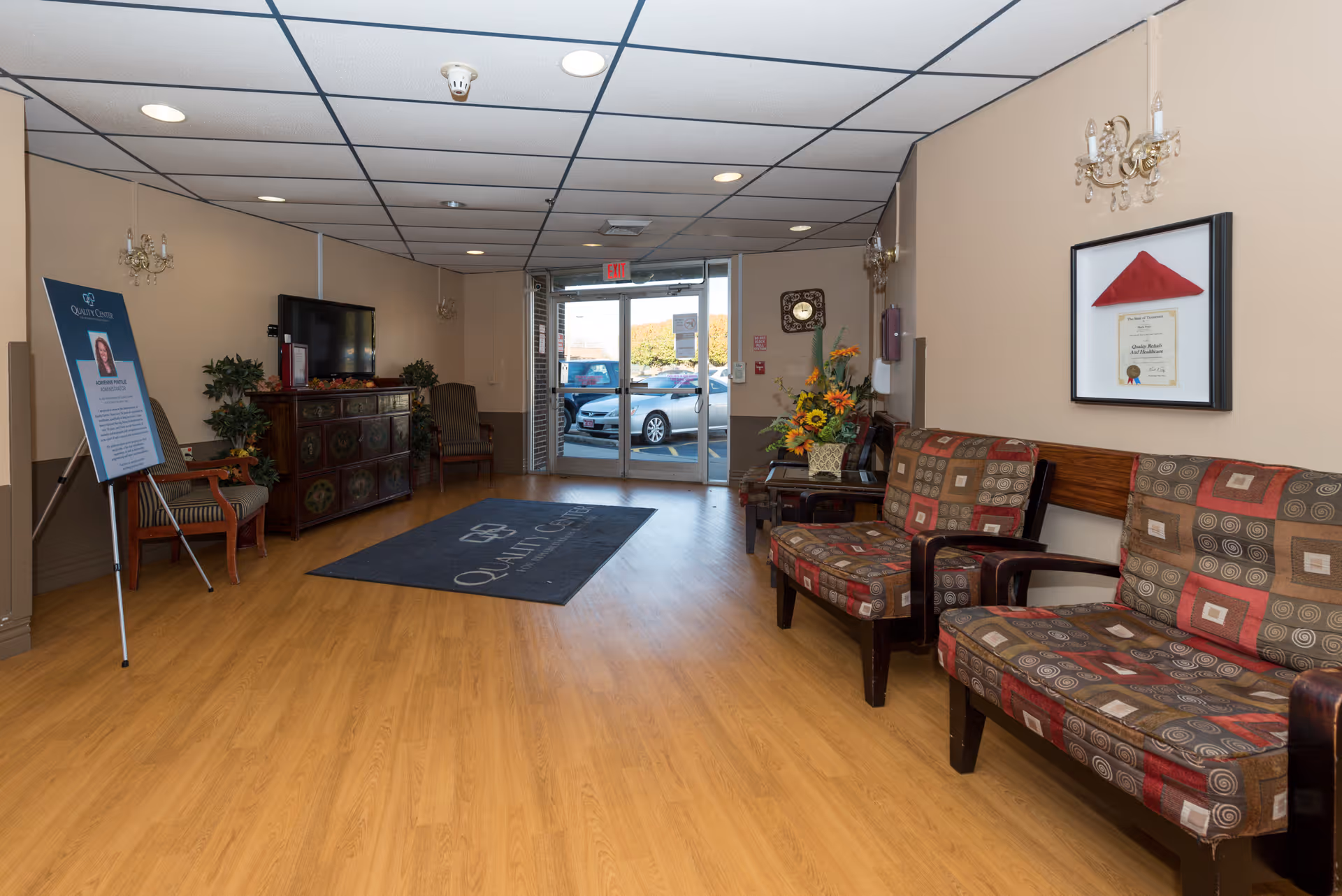 Interior view of a facility lobby with wooden flooring, patterned cushioned chairs along the right wall, a decorative cabinet with a TV on the left, a plant, a standing sign, and glass double doors leading outside with parked cars visible.