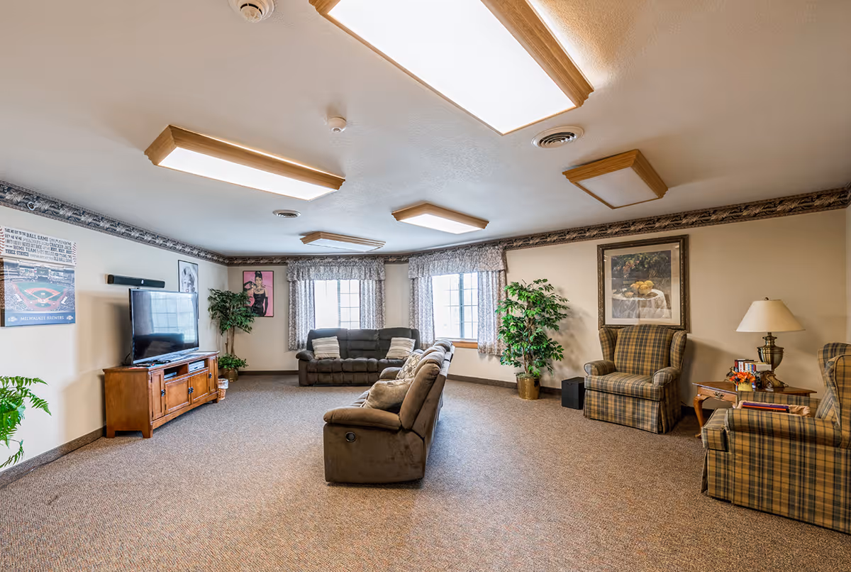 A spacious living room with beige walls and carpeted floor. The room features a large flat-screen TV on a wooden stand, two large windows with floral curtains, two brown sofas, two plaid armchairs, a side table with a lamp and flowers, framed artwork on the walls, and several potted plants.