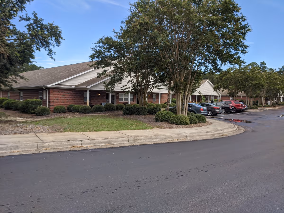 Exterior view of a single-story brick building with a sloped roof, surrounded by neatly trimmed bushes and trees. Several cars are parked along the driveway in front of the building under a clear blue sky.