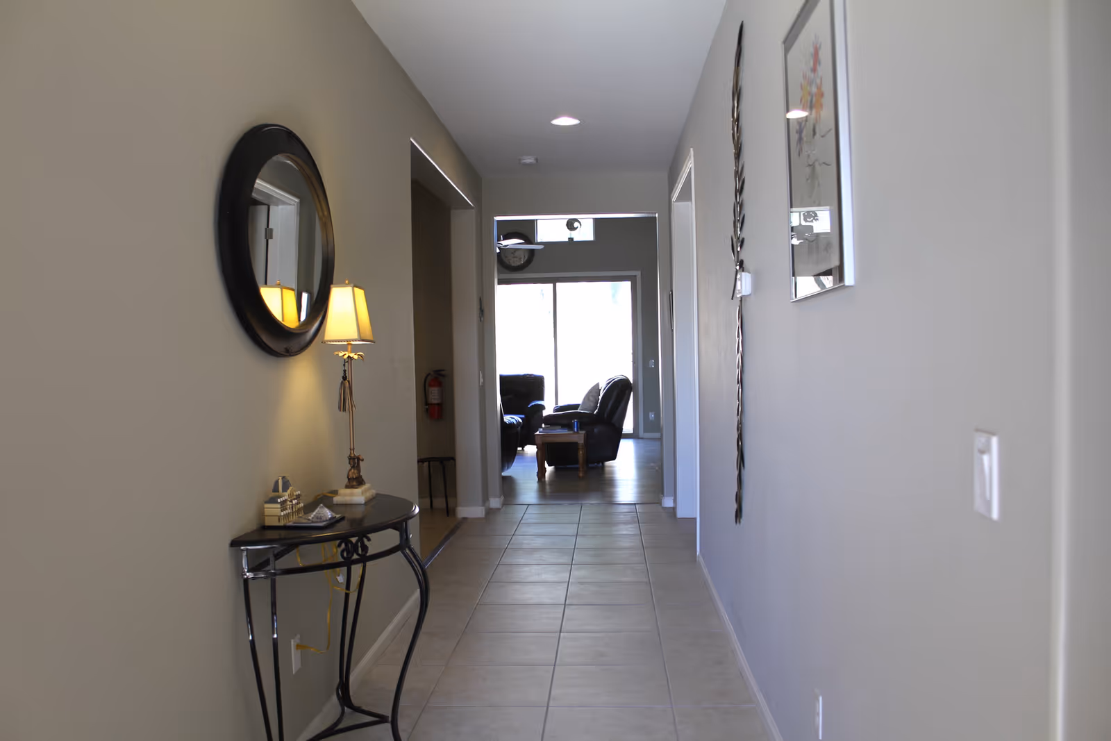 A hallway with beige tiled floor and light gray walls leading to a living room area with black leather chairs and a wooden coffee table. On the left side of the hallway, there is a small black table with a lamp and decorative items beneath a round mirror. On the right wall, there is a framed picture and a decorative wall piece.