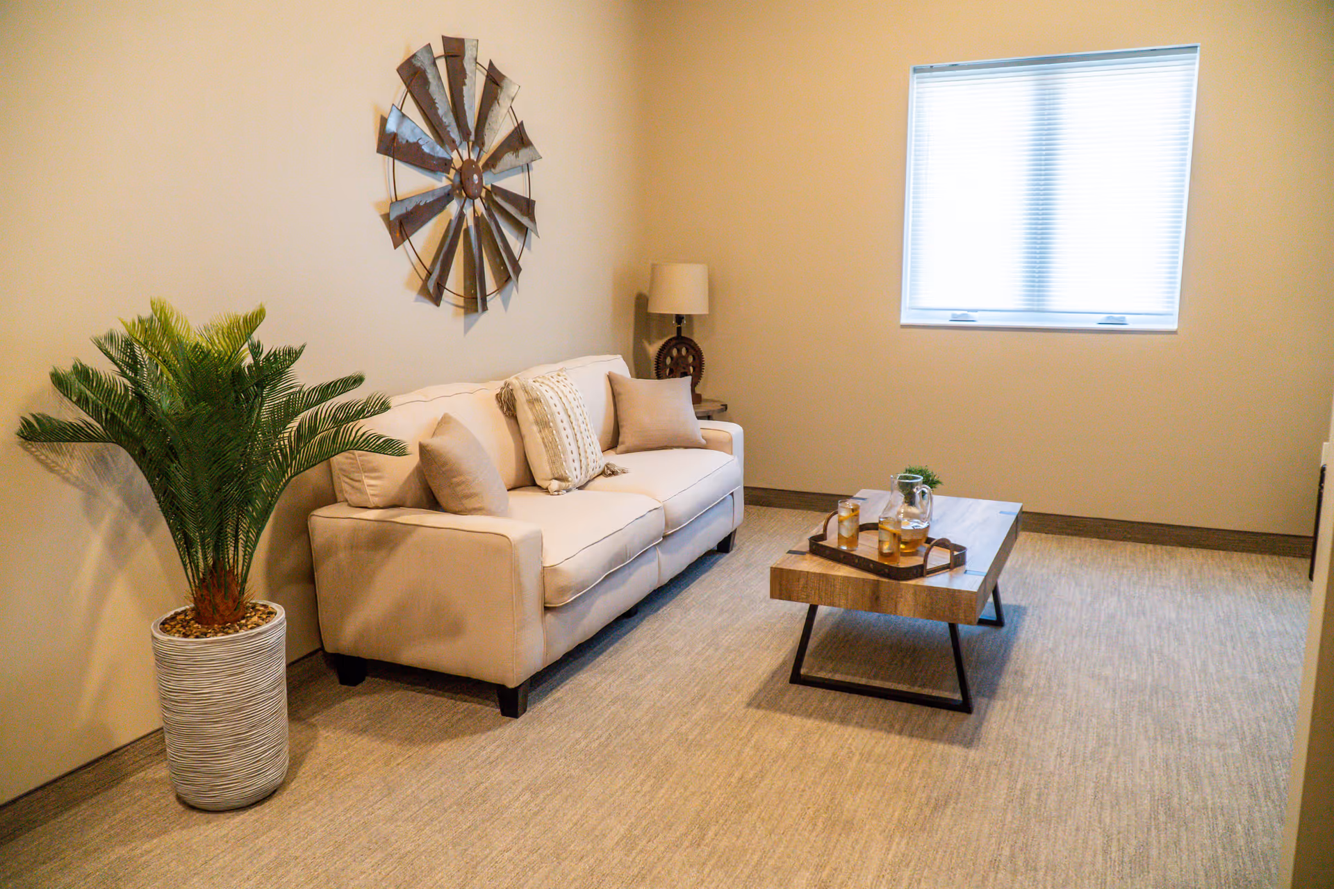 A cozy living room with a beige sofa adorned with cushions, a wooden coffee table with a tray holding a pitcher and glasses, a large potted plant in a white textured pot, a decorative metal wall art resembling a windmill, a table lamp on a side table, and a window with closed blinds letting in soft natural light.
