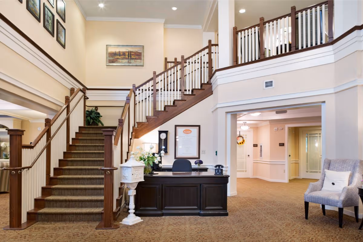 Interior view of a senior living facility lobby area with a carpeted staircase featuring wooden handrails and white balusters. There is a dark wooden reception desk with a chair, a vintage clock, and decorative flowers. The walls are painted beige with framed artwork and a chair with a cushion is placed to the right side. The area is well-lit with ceiling lights.