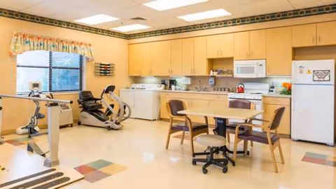 A bright senior living facility kitchenette and dining area with surrounding cabinetry and exercise equipment.