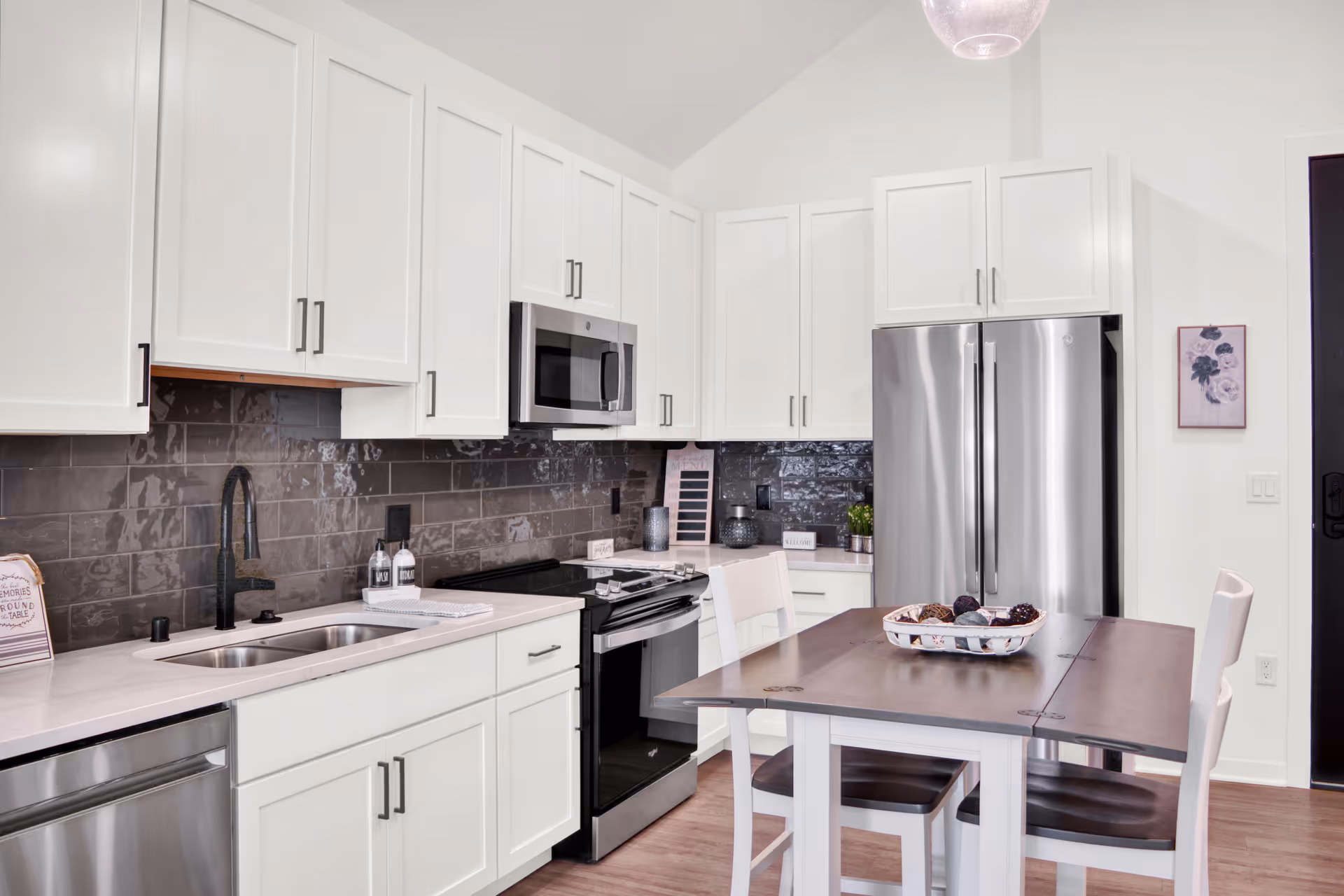 Modern kitchen with white cabinets, stainless steel appliances including a refrigerator, microwave, and dishwasher. There is a dark tile backsplash and a wooden dining table with four chairs in the center. The kitchen has a clean and bright appearance with a high ceiling and a hanging light fixture.