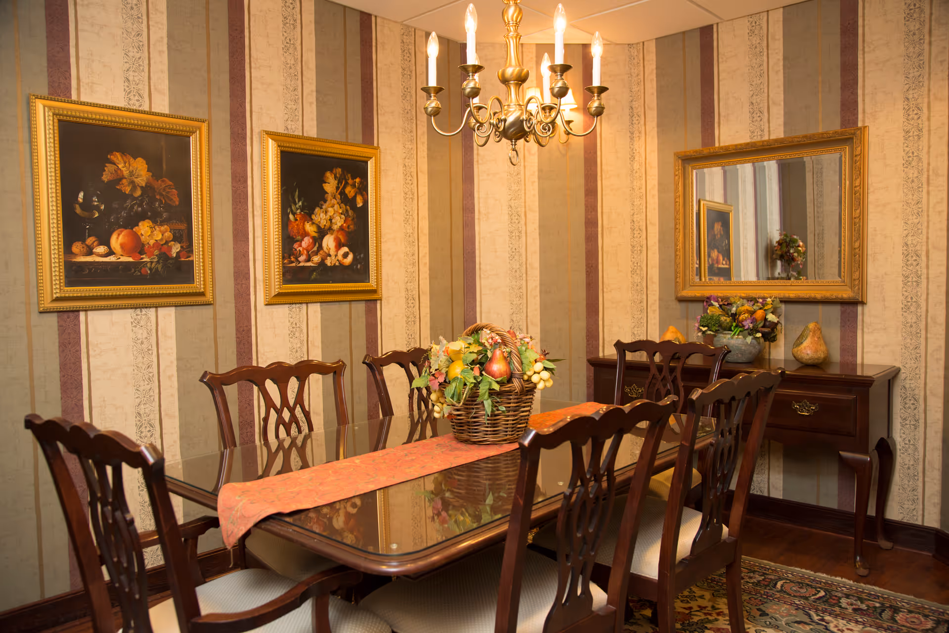 A traditional dining room with a glass-top wooden table surrounded by six wooden chairs with cushioned seats. A basket with artificial flowers and fruits is placed on a red table runner in the center of the table. The walls have vertical striped wallpaper in beige, brown, and purple tones. Two framed paintings of fruit still lifes hang on one wall, and a large rectangular mirror with a gold frame hangs on another wall above a wooden sideboard decorated with a floral arrangement and two decorative gourds. A brass chandelier with lit bulbs hangs above the table.