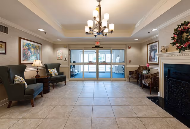 A spacious and well-lit senior living facility lobby with tiled flooring, two green armchairs with pillows on the left, two patterned armchairs on the right, a fireplace with a floral wreath above it, framed artwork on the walls, a chandelier hanging from the ceiling, and glass double doors leading outside.