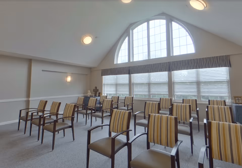 A room with multiple rows of striped cushioned chairs arranged facing forward. The room has a high ceiling with two round ceiling lights and a large arched window with multiple panes letting in natural light. There are a few decorative items on a cabinet against the back wall.