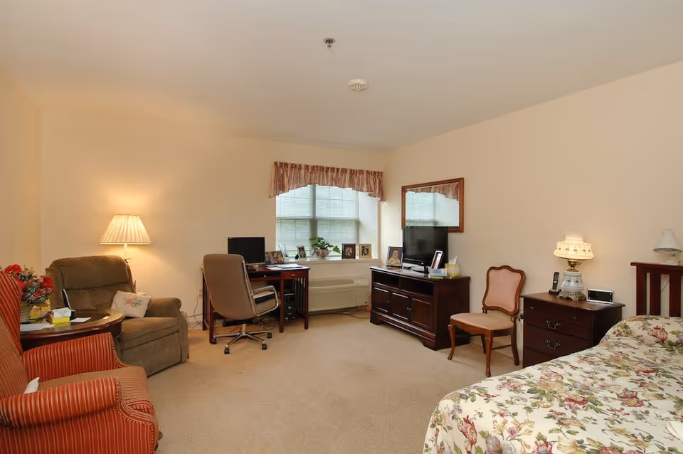 A cozy bedroom with beige walls and carpet. The room features a bed with floral bedding, a wooden nightstand with a decorative lamp, a wooden dresser with a flat-screen TV and a mirror above it, a wooden chair, a desk with a computer, and two armchairs with a small round table between them. A window with blinds and a valance lets in natural light.