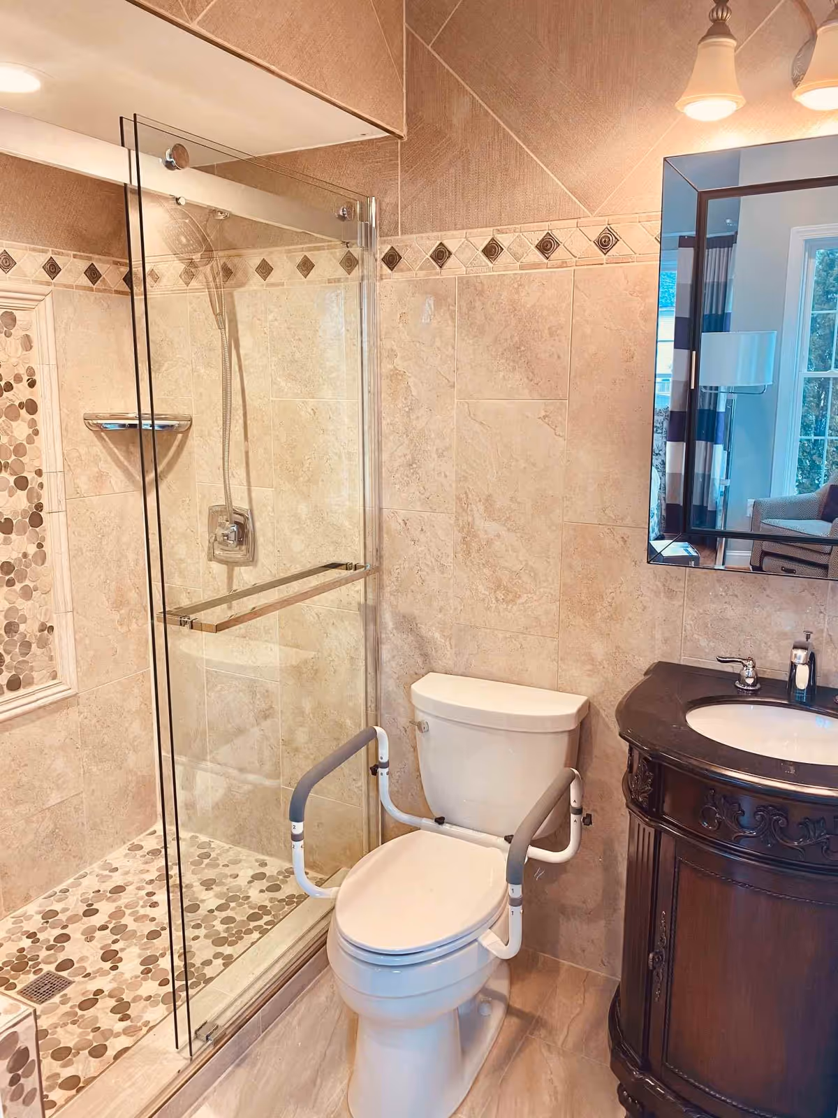 A bathroom with beige tiled walls and floor, featuring a glass-enclosed shower with pebble-style floor tiles and a handheld showerhead. There is a white toilet equipped with safety rails on both sides. Next to the toilet is a dark wooden vanity with a black countertop and a sink. A mirror above the vanity reflects a window and part of a living area with a chair and lamp.