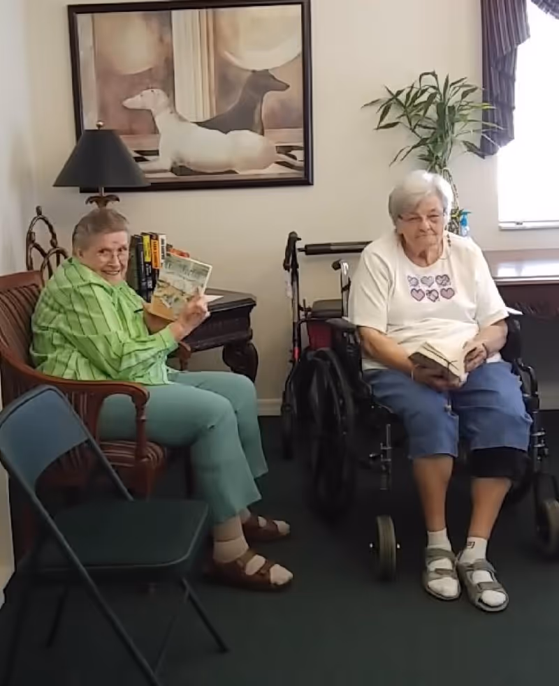 Two elderly women sitting indoors; one woman in a green outfit is seated on a wooden chair holding a book and smiling, while the other woman in a white shirt and blue shorts is seated in a wheelchair holding a book. Behind them is a side table with books and a lamp, a framed painting of two dogs on the wall, a potted plant, and a window with curtains.