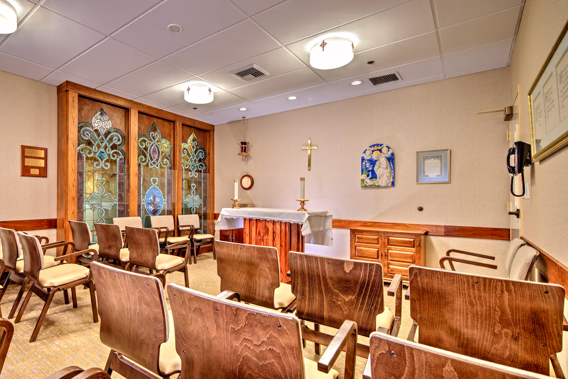 Small chapel room with wooden chairs arranged in rows facing an altar covered with a white cloth. The room features stained glass windows with decorative patterns, a wall-mounted cross, religious artwork, and soft ceiling lights.