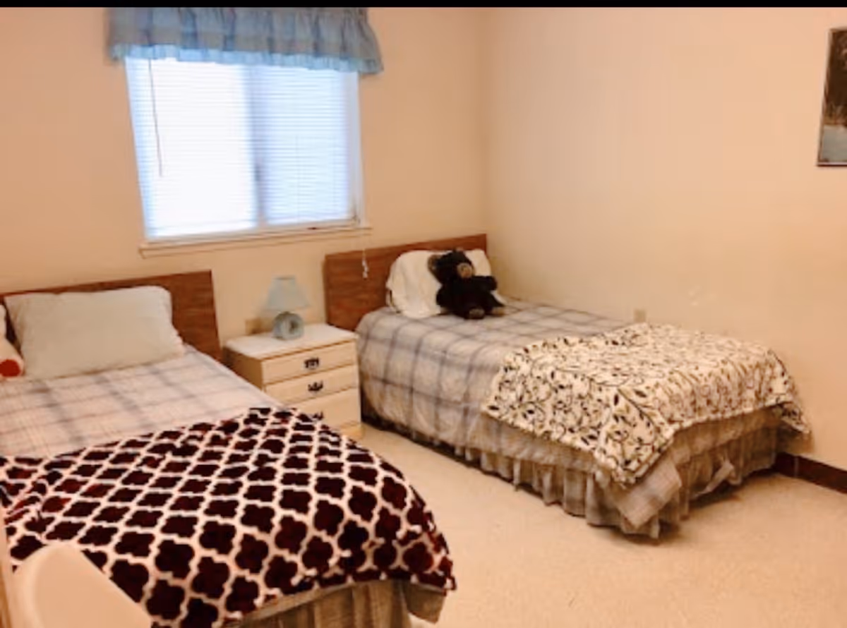 A bedroom with two single beds separated by a small white nightstand with a lamp. Each bed has a plaid bedspread and a decorative blanket at the foot. One bed has a black teddy bear on the pillow. There is a window with blinds and a blue valance above the nightstand.