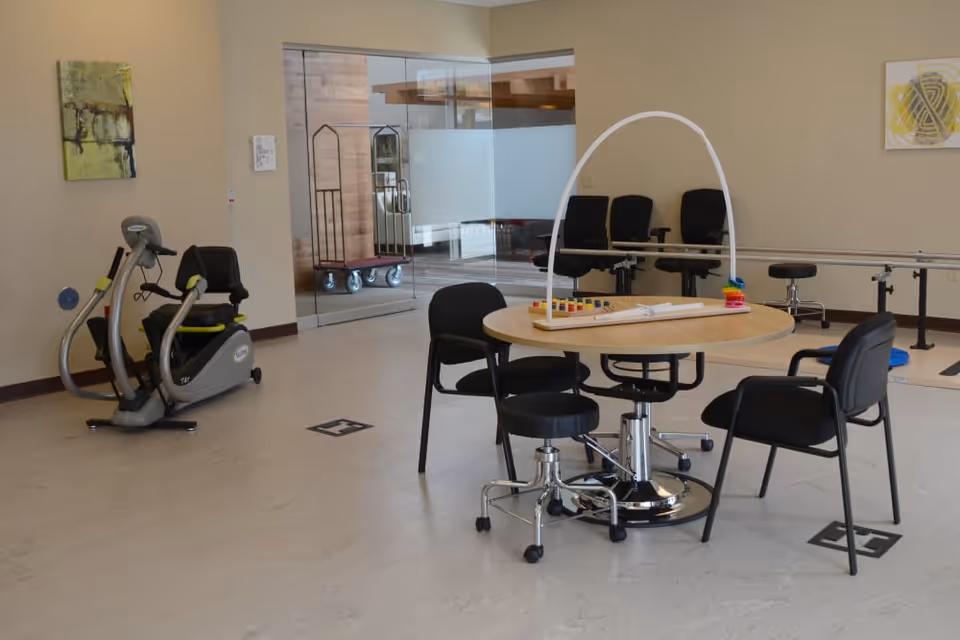 A therapy or activity room with a round table surrounded by four chairs, a therapy exercise bike, and parallel bars for walking assistance. The room has beige walls, a glass door, and some abstract artwork on the walls.