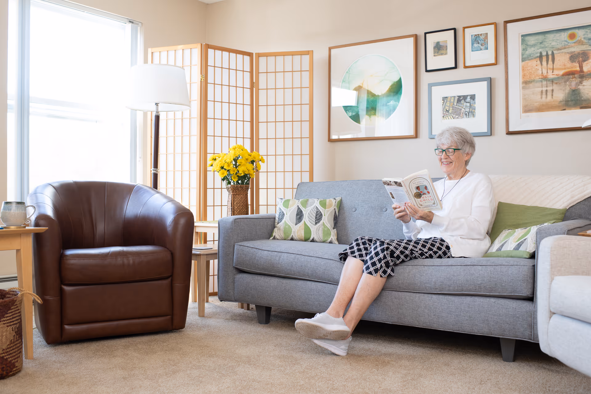 An elderly woman with short gray hair and glasses sits on a gray sofa in a bright living room, smiling while reading a book. The room features a brown leather armchair, a wooden side table with a mug, a floor lamp, a vase with yellow flowers, a folding screen, and several framed artworks on the wall.