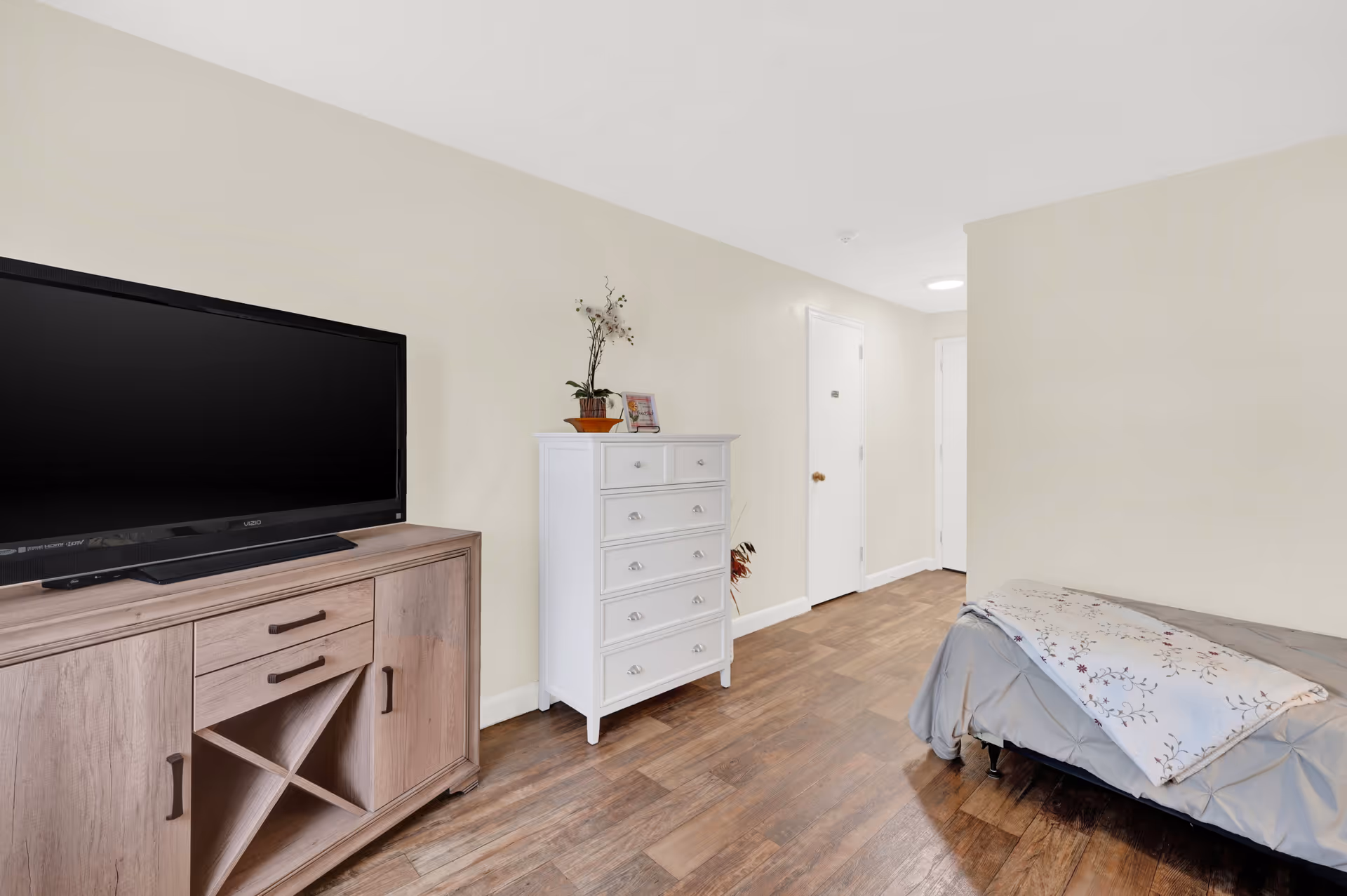 A simple bedroom with a wooden floor, a bed with gray bedding and a floral blanket, a white chest of drawers with a small plant and a picture frame on top, and a wooden TV stand with a large flat-screen television. The walls are light beige and there are two white doors visible in the background.