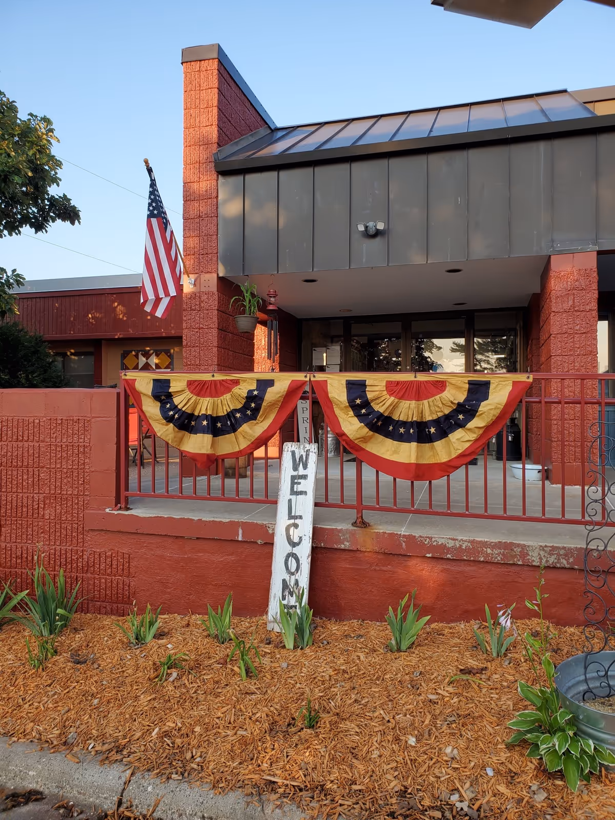 Entrance of a building with red brick walls and a metal roof. Two patriotic red, yellow, and blue bunting decorations hang on a red railing. An American flag is mounted on the left side of the entrance. A white wooden sign with the word 'WELCOME' written vertically is placed in a mulched flower bed with green plants in front of the railing.