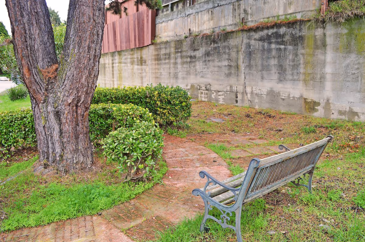 A small outdoor garden area featuring a curved brick pathway, a wooden bench with metal armrests, green bushes, a large tree, and a concrete wall in the background.