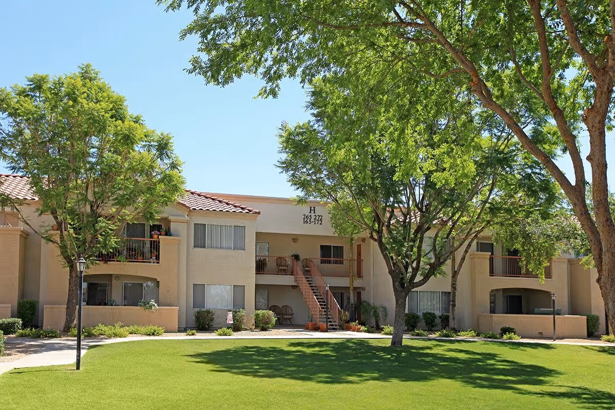 Exterior view of a two-story residential building with beige walls and red-tiled roof, surrounded by green trees and a well-maintained lawn under a clear blue sky.