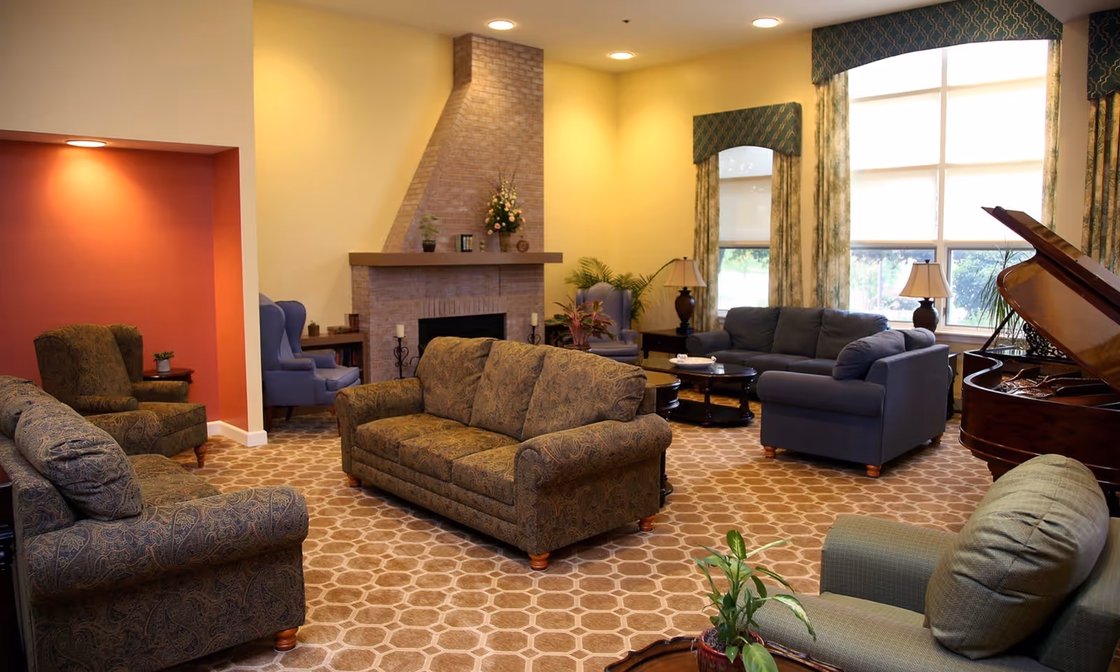 A cozy living room with patterned sofas and armchairs arranged around a brick fireplace. The room features large windows with decorative curtains, a grand piano, table lamps, and plants, creating a warm and inviting atmosphere.