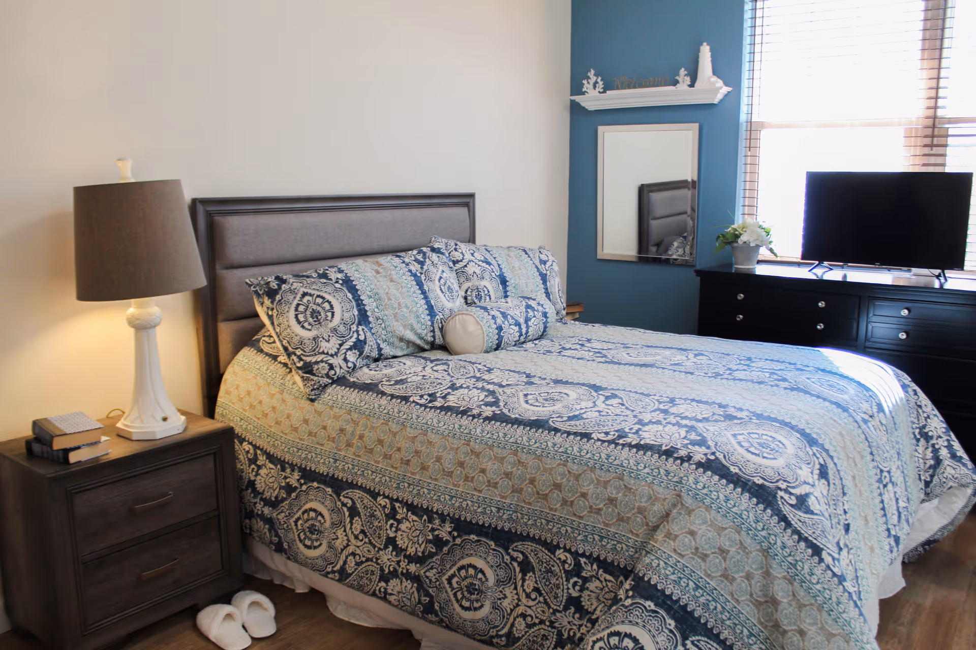 Bedroom featuring a blue-pattern quilt, nightstand with lamp, slippers, and a dresser with a TV.