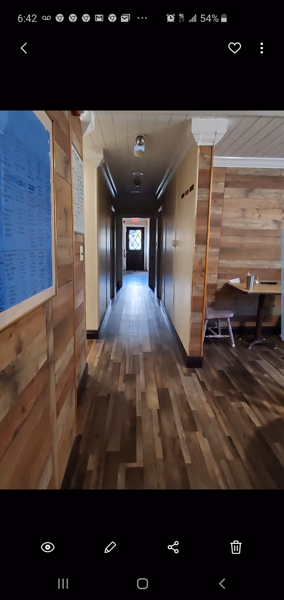 A long, narrow hallway with wooden flooring and beige walls. On the left side, there is a blue bulletin board mounted on a wooden wall. On the right side, part of a small table and chair are visible against a wooden paneled wall. At the end of the hallway, a door with a window is visible, letting in some natural light.