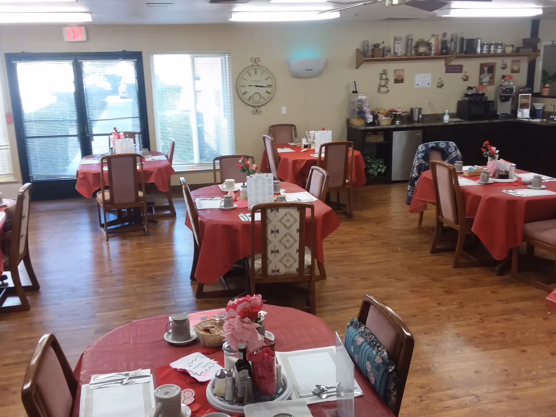 Dining room with multiple round tables covered with red tablecloths, each set with cups, plates, utensils, and floral centerpieces. Wooden chairs surround the tables, and the room has wooden flooring. In the background, there is a counter area with coffee machines and other kitchen items, a large clock on the wall, and windows letting in natural light.