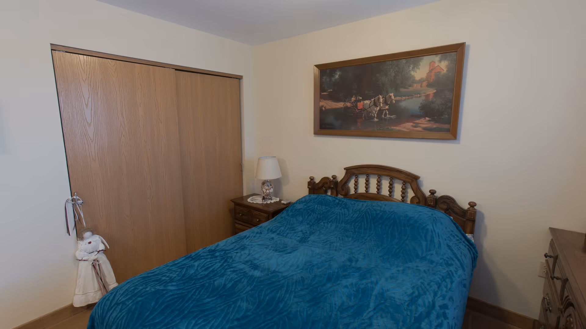 A bedroom with a wooden bed frame covered by a blue blanket. Next to the bed is a wooden nightstand with a lamp and some small items. A large framed painting depicting a horse-drawn carriage near a river hangs on the wall above the bed. There is a wooden closet with sliding doors and a decorative doll hanging on the wall beside it.