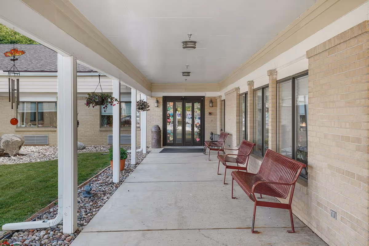 Covered outdoor walkway with hanging flower pots and red metal benches along a beige brick wall, leading to a glass door entrance decorated with colorful paper cutouts.