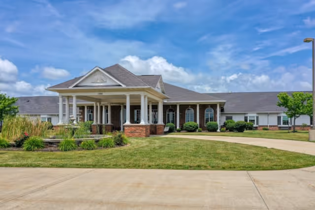 Front exterior view of Cedarhurst Senior Living of Godfrey building with a covered entrance, brick and white siding, surrounded by green lawn and landscaping under a partly cloudy blue sky.
