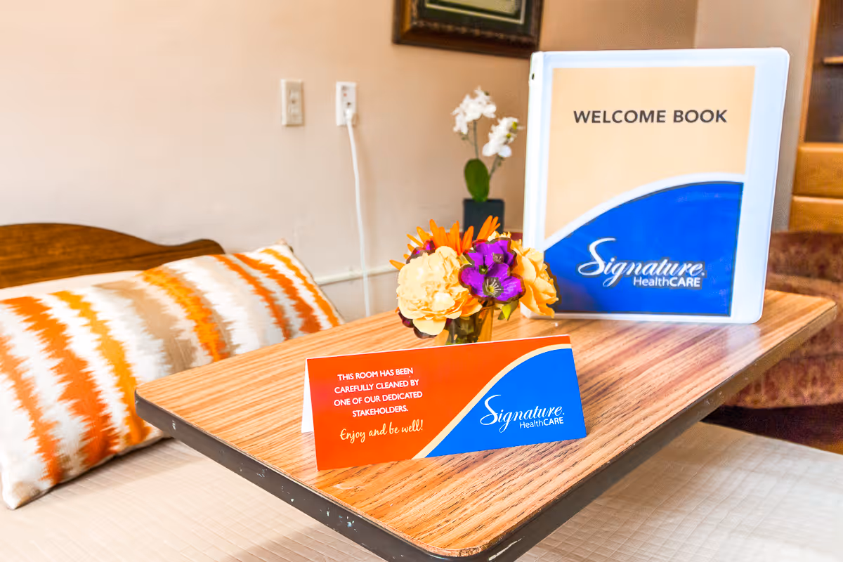 A wooden overbed table with a small bouquet of colorful flowers, a welcome book labeled 'Signature HealthCARE', and a card stating the room has been carefully cleaned. The table is positioned over a bed with a white and orange patterned pillow and a beige bedspread. The background shows a beige wall with electrical outlets and a framed picture.