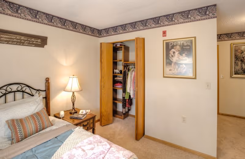 A cozy bedroom with a bed featuring multiple pillows and a patterned blanket. A wooden nightstand with a decorative lamp, a candle, and some books sits next to the bed. An open closet with wooden bi-fold doors reveals hanging clothes and shelves. The walls are light-colored with a decorative border near the ceiling, and framed artwork is hung on the walls.