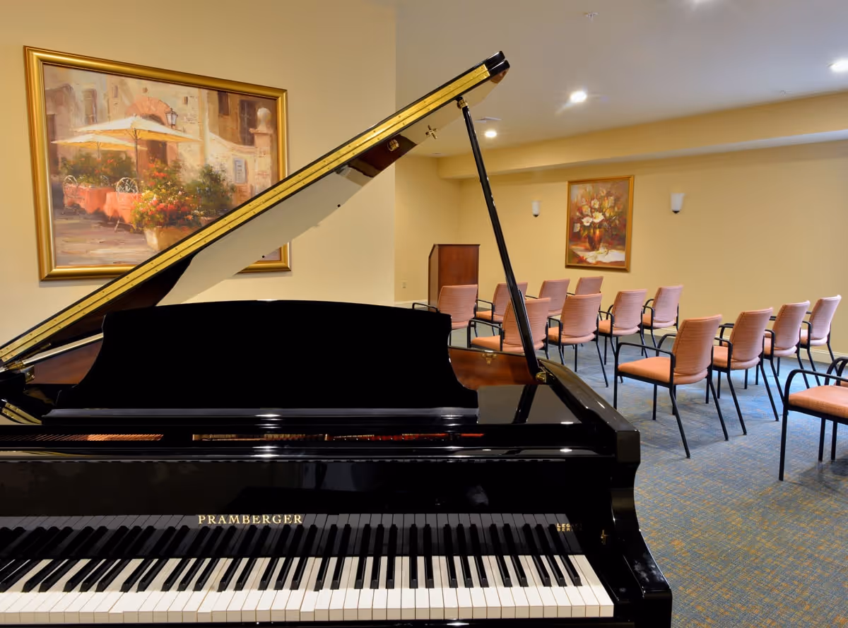 Interior room with a black grand piano in the foreground and rows of pink cushioned chairs arranged facing a wooden podium. The walls are beige with two framed paintings, one depicting an outdoor cafe scene and the other a floral arrangement.